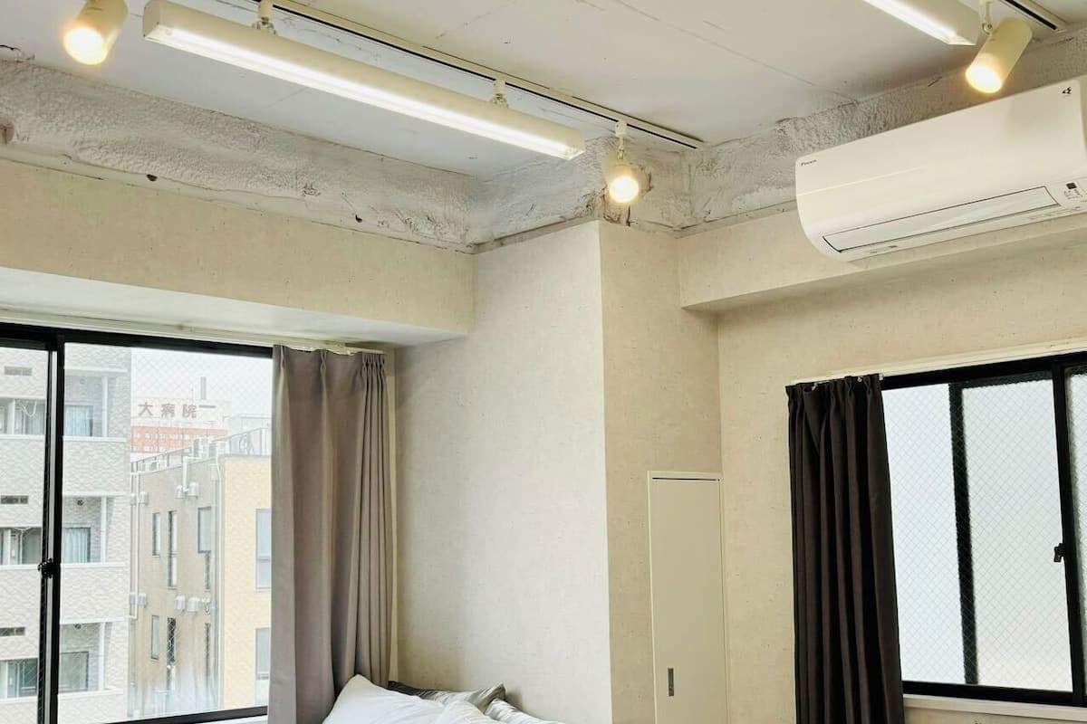 A hotel room with two beds, a window with curtains, an air conditioning unit, and a view of buildings outside.