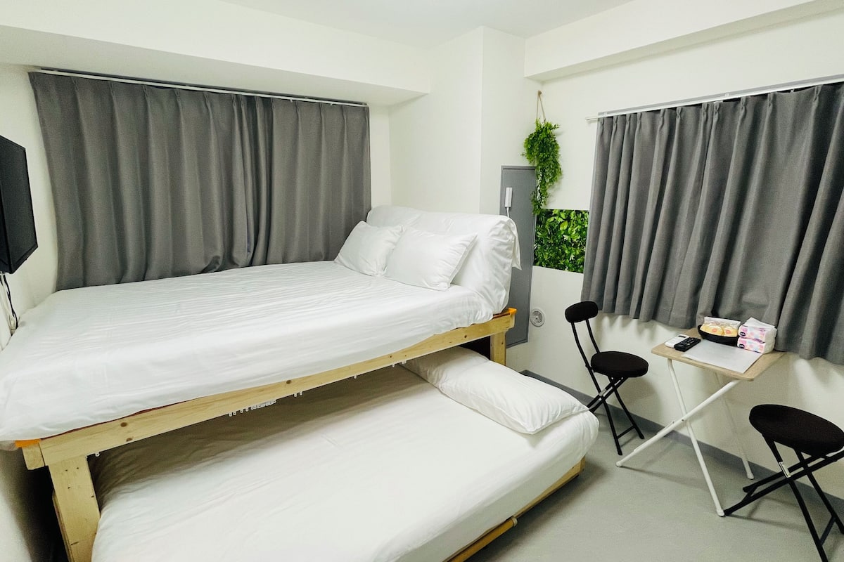 A small, single-room accommodation with a bunk bed, a desk, two chairs, and a small window with curtains.