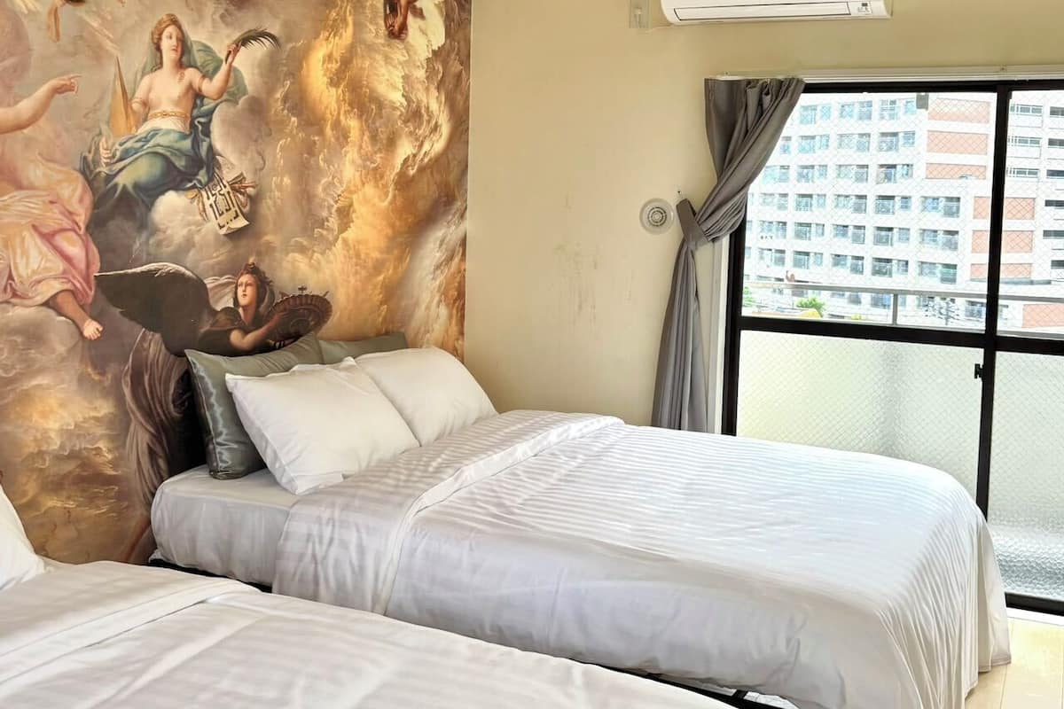 A hotel room with two beds, a large mural on the wall, an air conditioning unit, and a view of a building through a window.