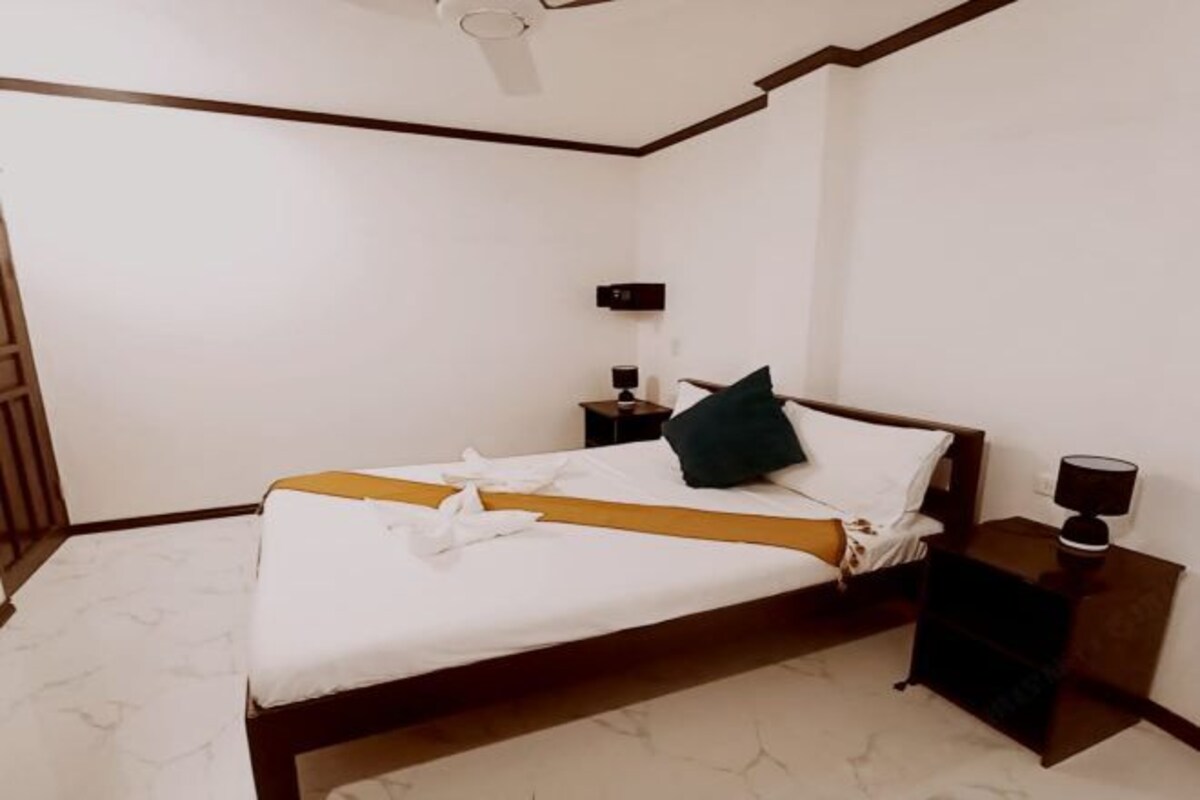 Una cama bien tendida con una colcha blanca y amarilla, un almohadón verde oscuro y una mesita de noche con una lámpara.
