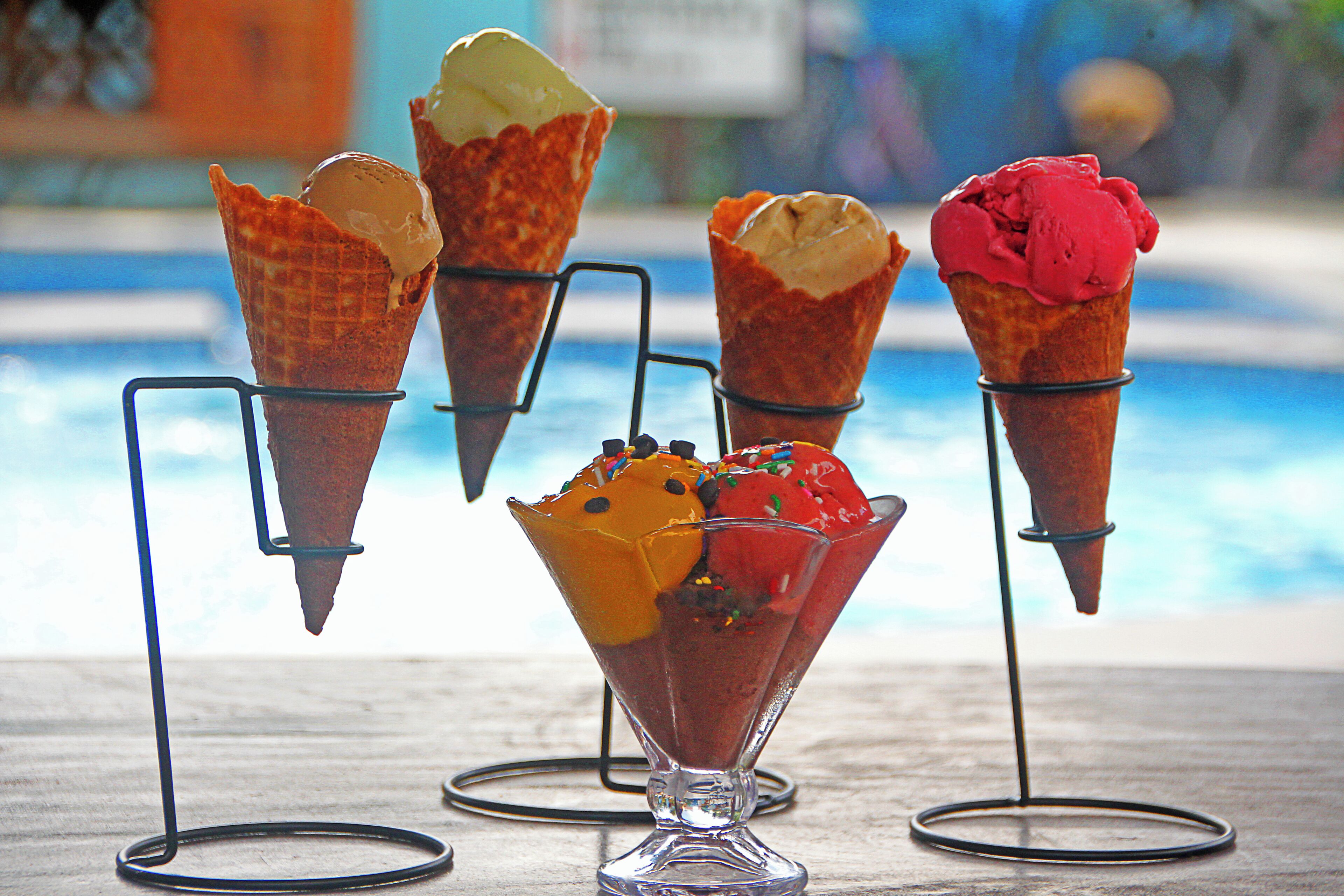 Quatre cornets de glace et une coupe glacée sur une table, avec une piscine en arrière-plan.