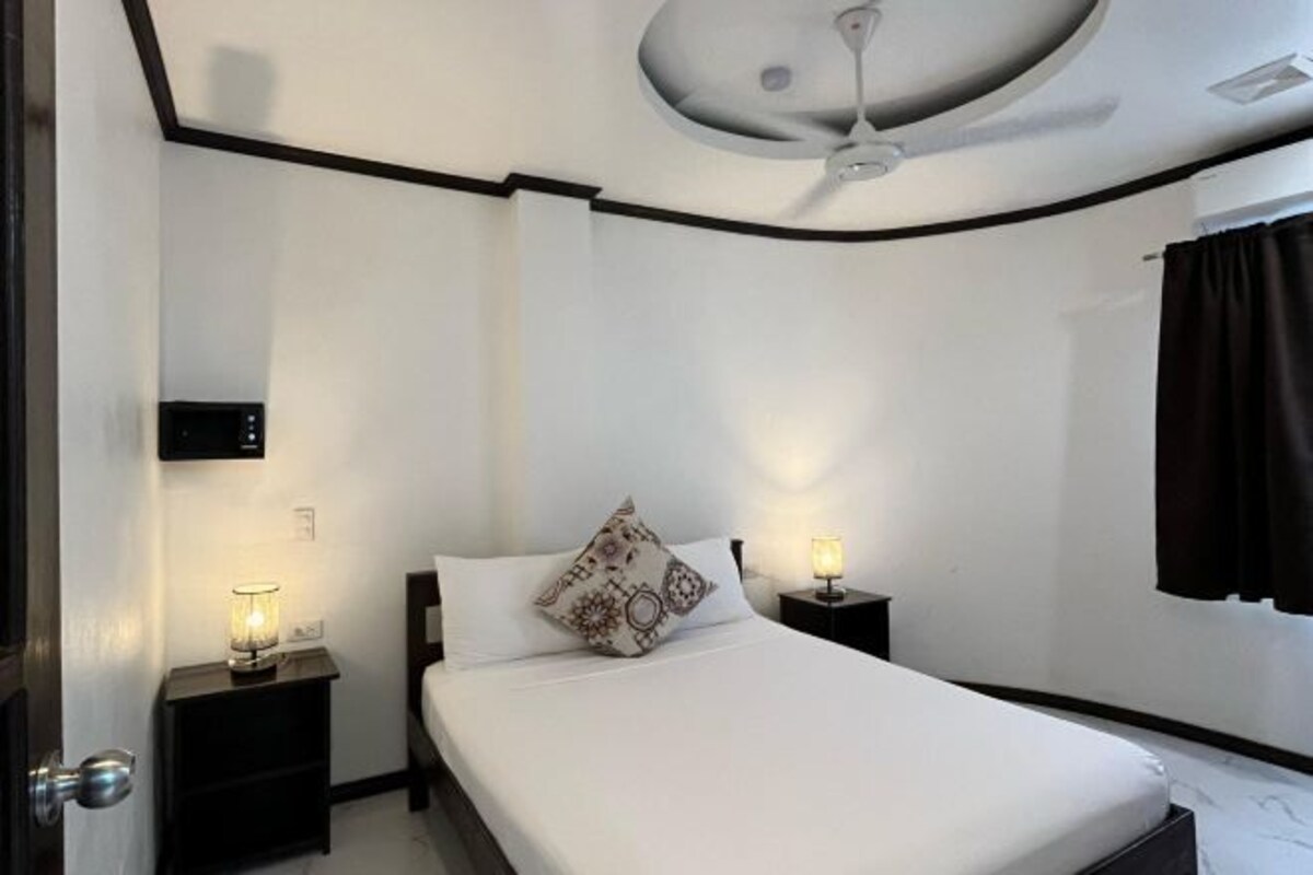 Una camera da letto con un letto, due comodini con lampade, una ventola a soffitto e una finestra con tende.