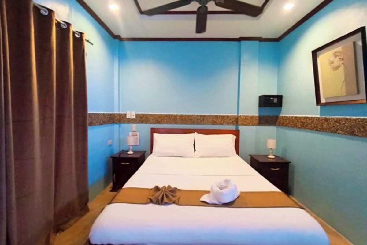 Una camera d'albergo con un letto, due comodini, una ventola a soffitto e un quadro appeso al muro.