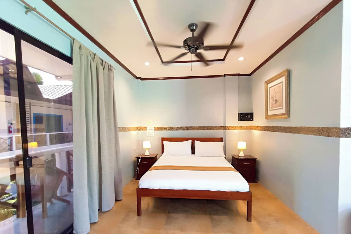 A hotel room with a large bed, two bedside tables with lamps, a ceiling fan, a window with curtains, and a framed picture on the wall.
