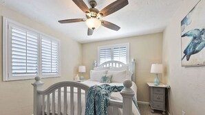 Quarto com cama branca, ventilador de teto, duas mesas de cabeceira com luminárias e janela com persianas.