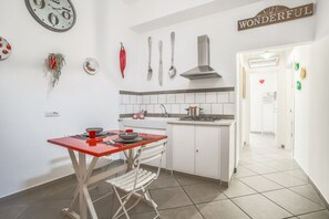 Una cocina compacta con armarios blancos, una mesa roja y una silla de comedor. Incluye detalles decorativos en la pared como un reloj, utensilios y un letrero que dice "LIFE IS WONDERFUL".