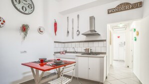 A compact kitchen with white cabinets, a red table, and a dining chair. There are wall decorations including a clock, utensils, and a sign that says "LIFE IS WONDERFUL". - 4 Bedrooms Apt in Sorrento Center | location&view (Sorrento)