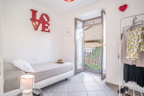 Un dormitorio pequeño y moderno con una cama individual, una mesita de noche con lámpara, un letrero rojo con la palabra 'LOVE' en la pared y un balcón con vistas a zonas verdes.