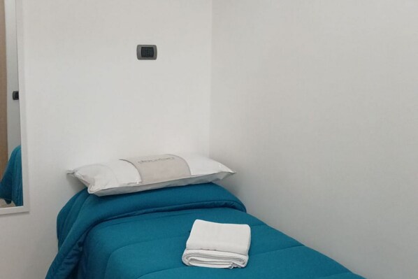 Een eenpersoonsbed met een blauwe matras en een wit kussen, een opgevouwen witte handdoek, en een klein bedieningspaneel aan de muur.