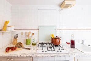 A kitchen with white tiles, a gas stove, a pot, a cutting board with tomatoes, a knife, and a bottle of red wine.
