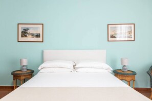 A neatly made bed with white linens and pillows, flanked by two wicker nightstands with lamps, against a light blue wall with two framed pictures.