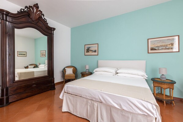 A bedroom with a large mirror, a bed with white linens, two nightstands with lamps, and two framed pictures on the wall.