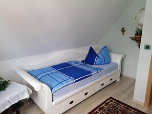 En vit säng med ett blått, rutigt sängöverkast och kuddar, ett litet bord med en spetsduk och en mönstrad matta på ett trägolv.