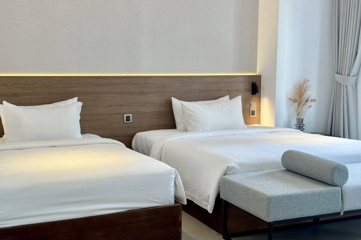 Una habitación de hotel con dos camas, un banco, una mesita de noche y una lámpara fijada a la pared.