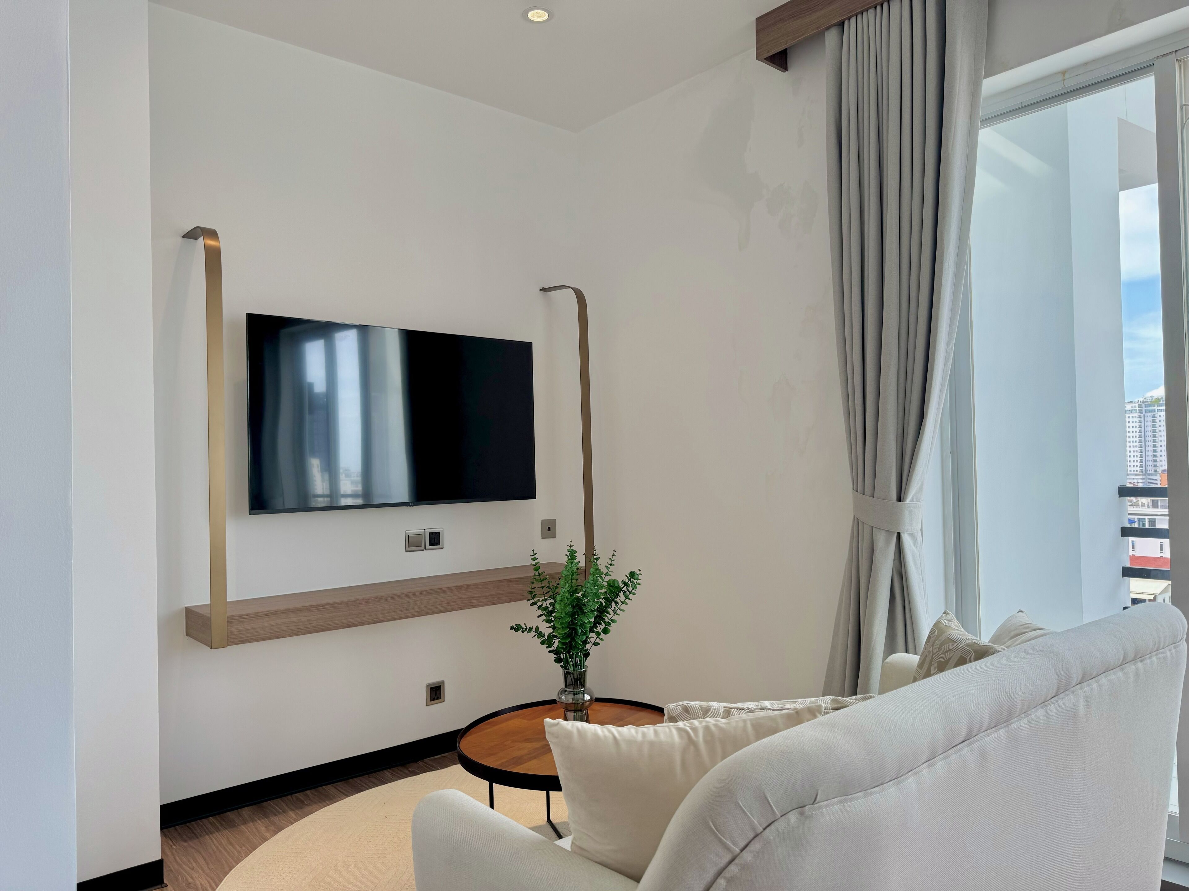 Uma sala de estar moderna com televisão de tela plana na parede, uma mesa redonda de madeira, um sofá branco e um vaso com planta.
