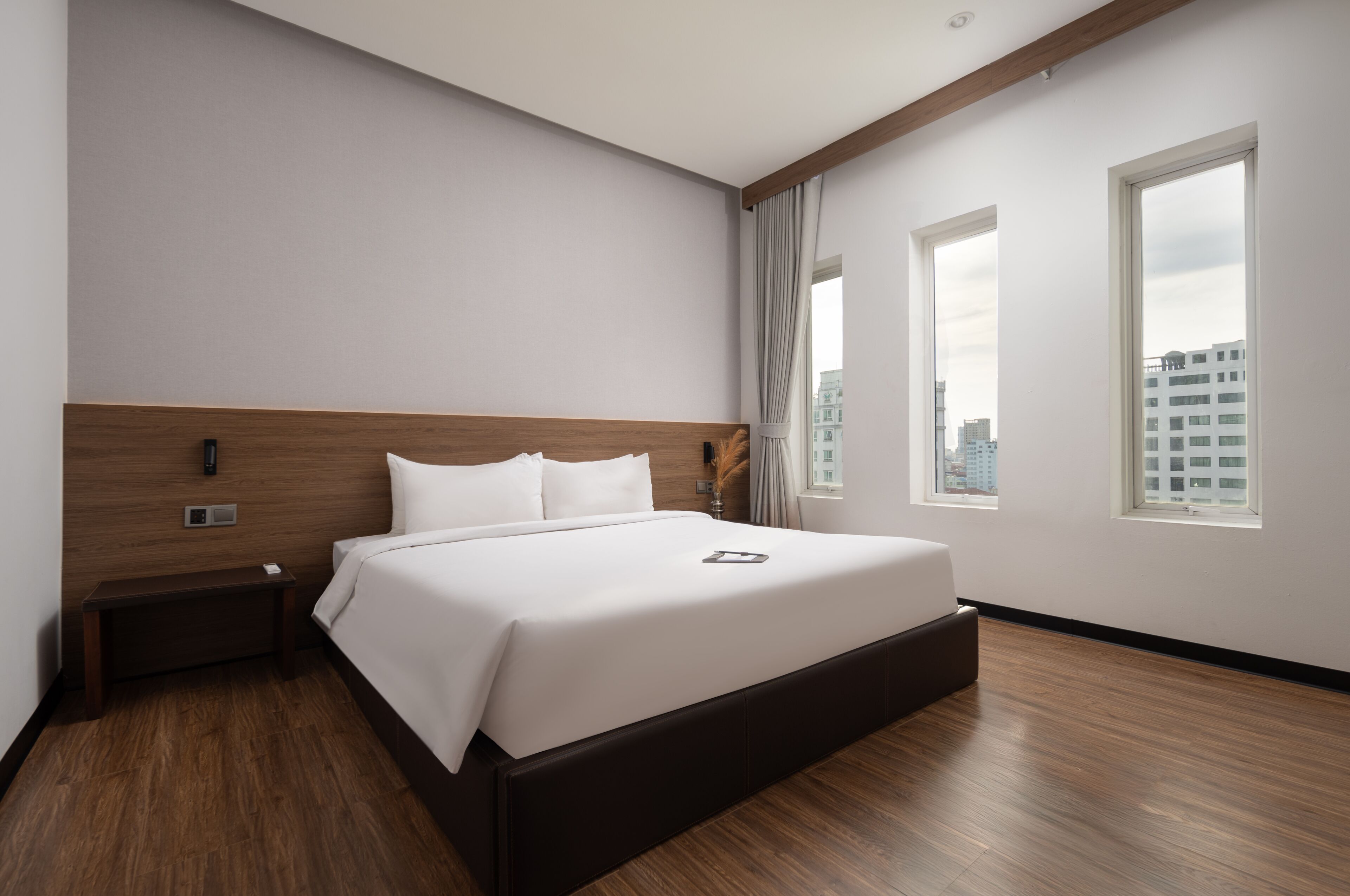 Een moderne hotelkamer met een groot bed, een houten hoofdbord, een nachtkastje en uitzicht op de stad door twee ramen.