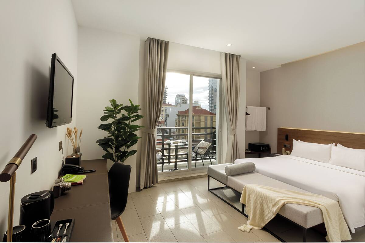 Habitación de hotel con cama, televisor, escritorio con planta y balcón con sillas, y vista a edificios.