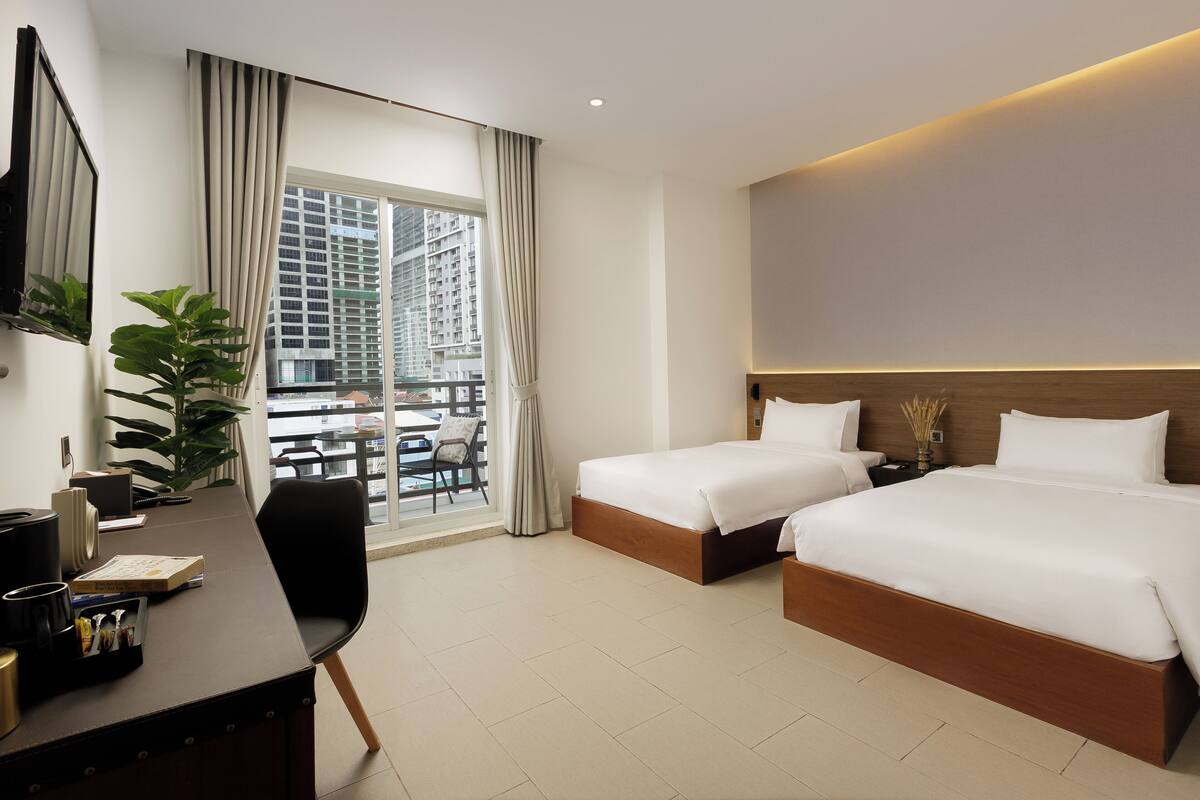 Habitación de hotel con dos camas, un escritorio, una silla y un ventanal con vistas a edificios altos.