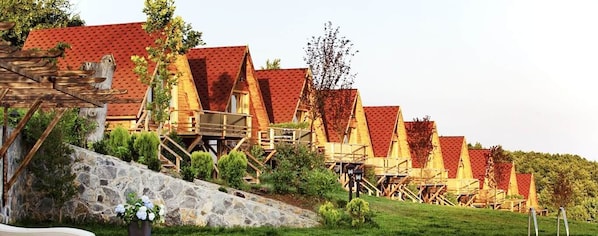 A row of wooden chalets with red roofs, elevated on stilts, surrounded by greenery and a stone wall. - Vadi Zirkon Otel (Karamürsel)