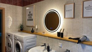 A modern bathroom with a washing machine, a sink, and a mirror with a circular light. There are framed signs on the wall, one of which reads "NO SELFIES IN THE BATHROOM." - Cozy apartment with conservatory near the safari park - dogs welcome (Verl)
