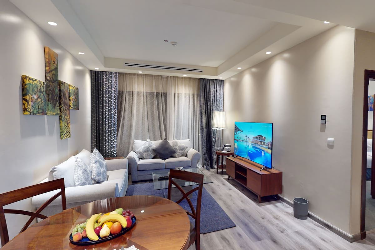 A modern living room with a sofa, a dining table with fruit, a TV, and a view of a building through the window.