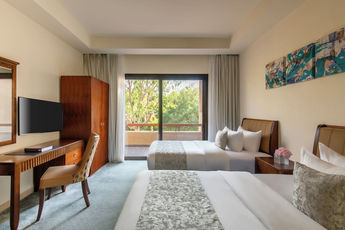 A hotel room with a large bed, a desk with a TV, and a balcony with a view of trees.