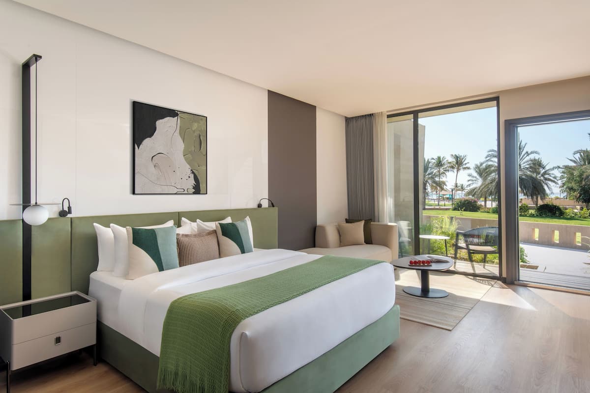 A modern hotel room with a large bed, a green headboard, a painting on the wall, and a balcony with a view of palm trees.
