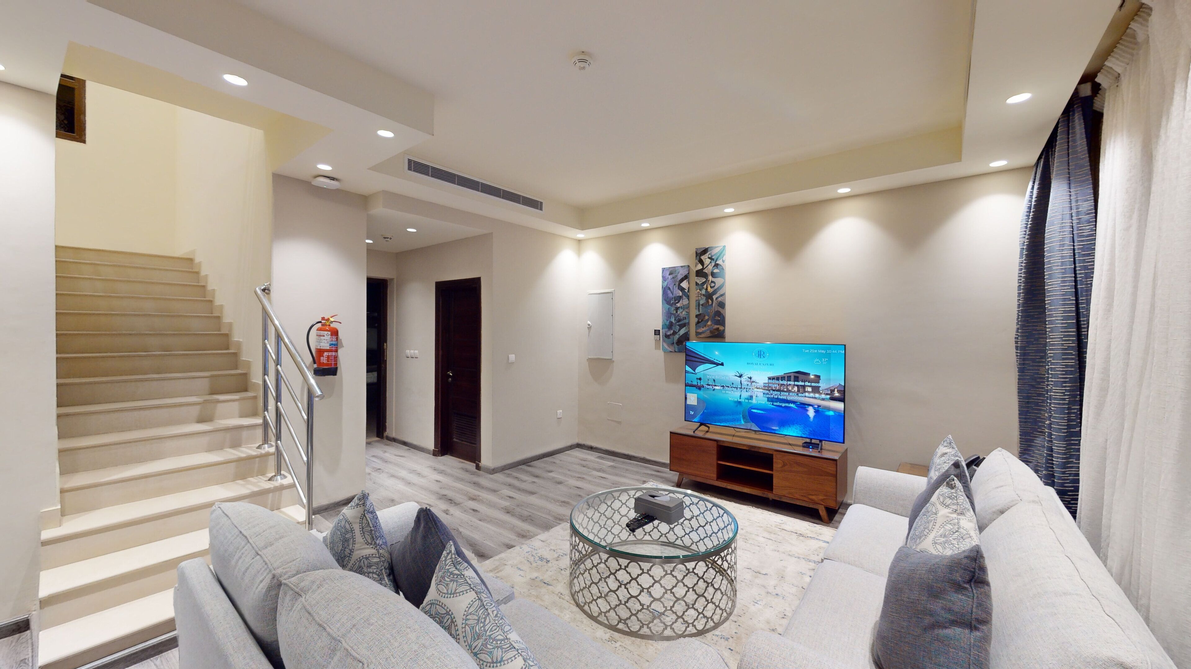 A modern living room with a staircase, a flat-screen TV, and a glass coffee table.