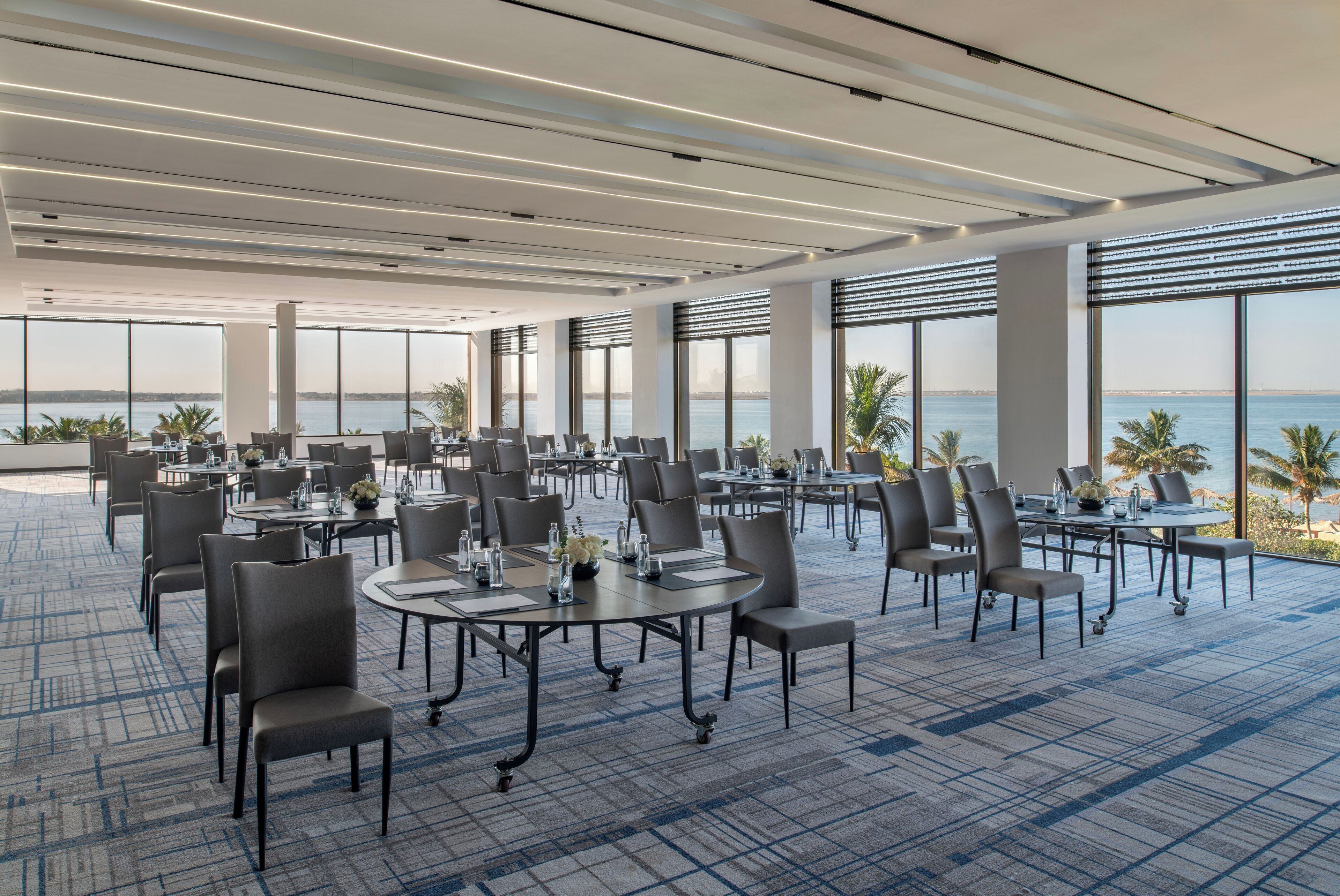 A spacious conference room with tables and chairs, large windows, and a view of the sea.
