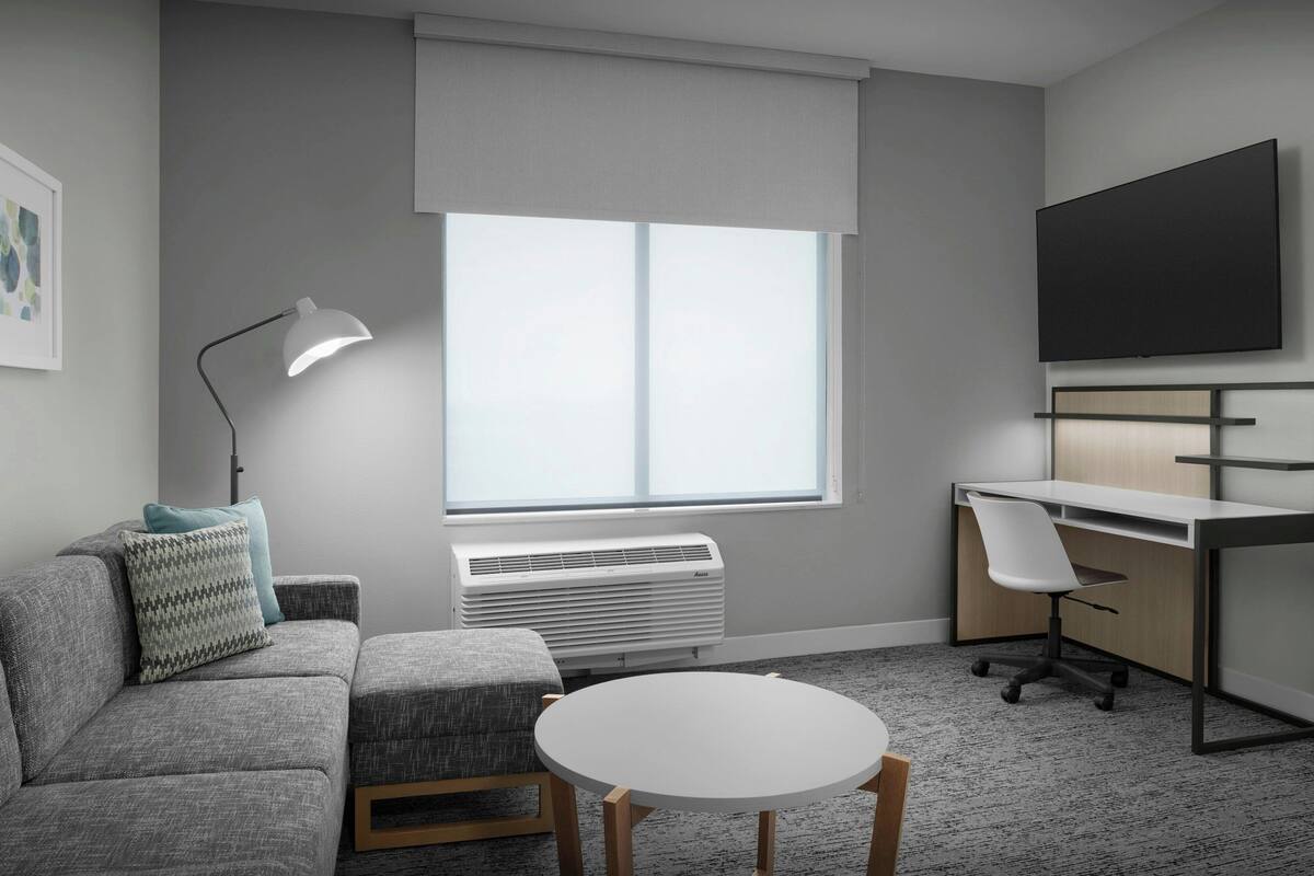 Een moderne kantoorruimte met een grijze bank, een witte tafel, een bureau, een stoel, een staande lamp en een groot raam met jaloezieën.