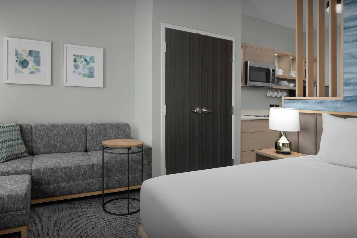 Een moderne hotelkamer met een grijze bank, een rond bijzettafeltje, een bed met wit beddengoed, een donker houten deur, een klein houten nachtkastje met een lamp, een magnetron en een kast met keukengerei.