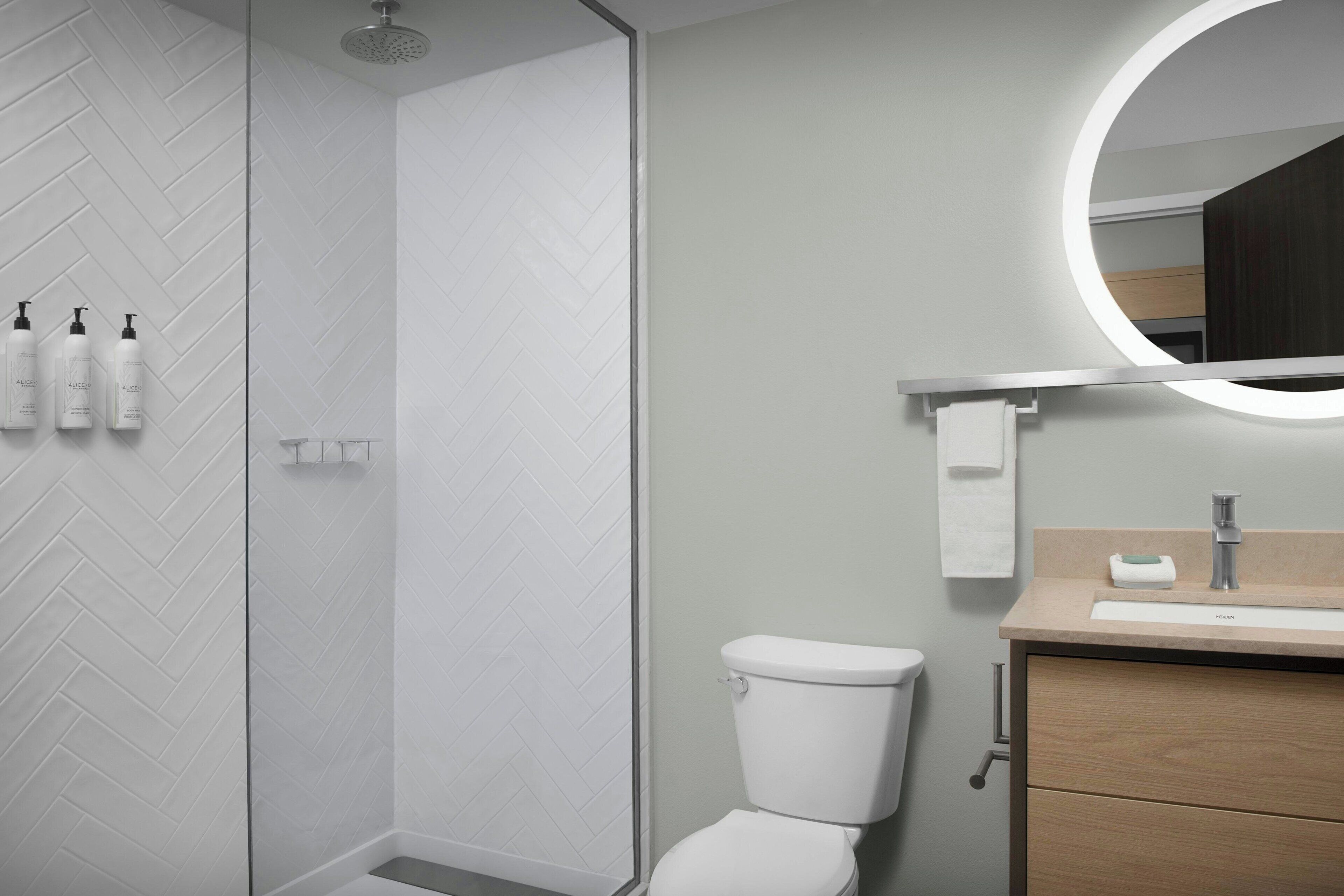 A modern bathroom with a herringbone tile wall, a shower, a toilet, a sink with a mirror, and toiletries on the wall.