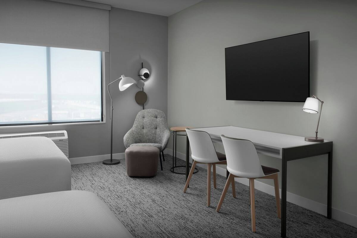 Een moderne hotelkamer met een groot raam, een bed, een bureau met twee stoelen, een rond poefje en een tv aan de muur.