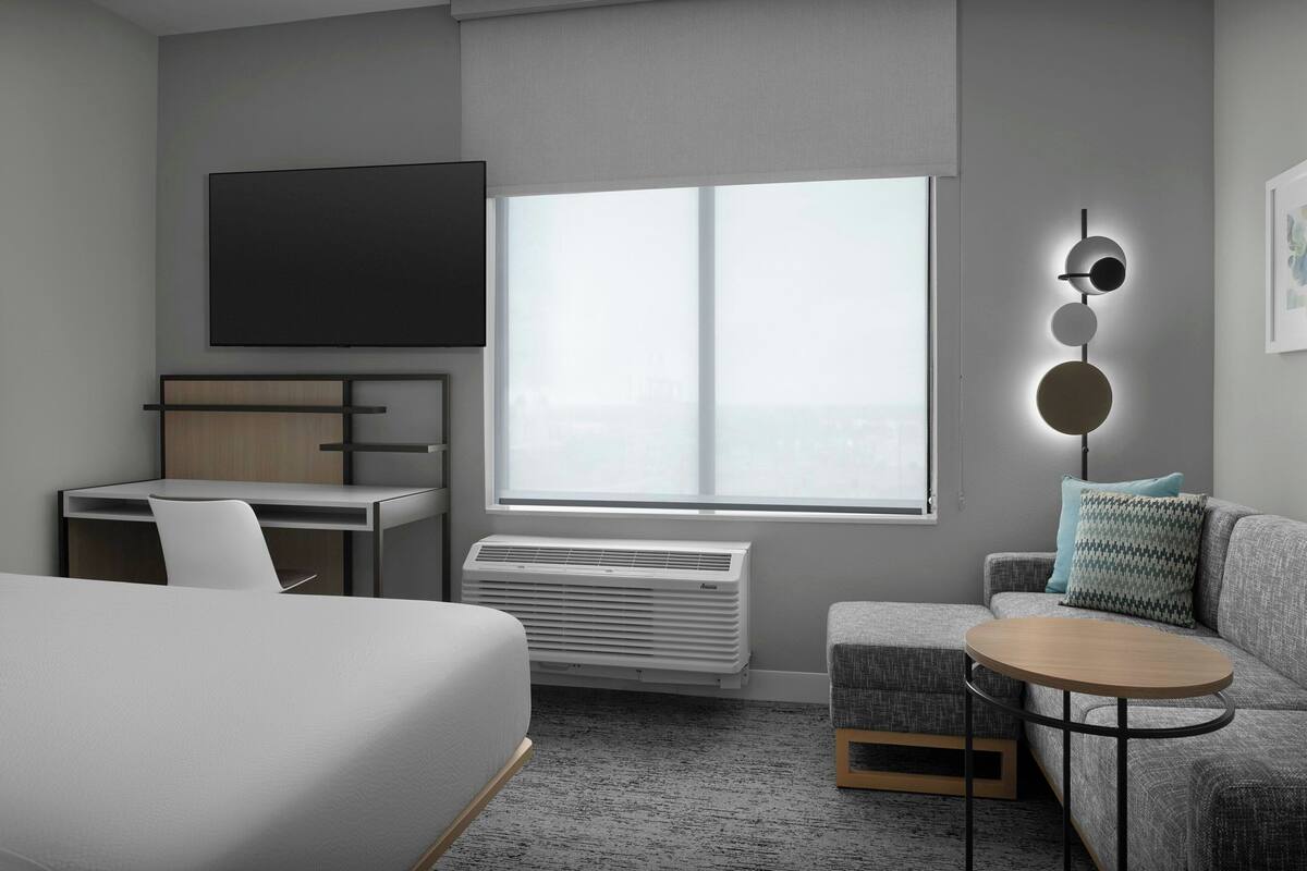 Een moderne hotelkamer met een bed, een bureau, een slaapbank, een ronde tafel, een televisie, een raam en een wandlamp.