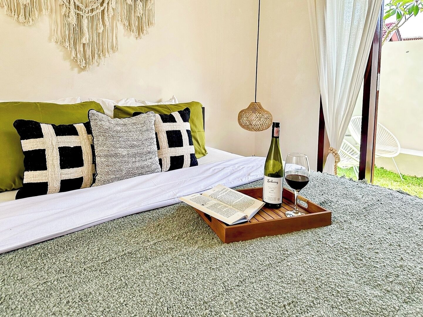 Ein ordentlich gemachtes Bett mit einem grün-weiß karierten Kissen, einer Weinflasche und einem Glas auf einem Tablett, im Hintergrund ein Wandbehang aus Makramee und eine Korbtasche-Lampe.