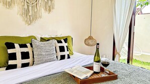 Ein ordentlich gemachtes Bett mit einem grün-weiß karierten Kissen, einer Weinflasche und einem Glas auf einem Tablett, im Hintergrund ein Wandbehang aus Makramee und eine Korbtasche-Lampe.