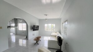 A spacious living room with a white sofa, a small table, and a TV mounted on the wall. There are windows allowing natural light and a ceiling fan. - The Retreat (Lyssons)