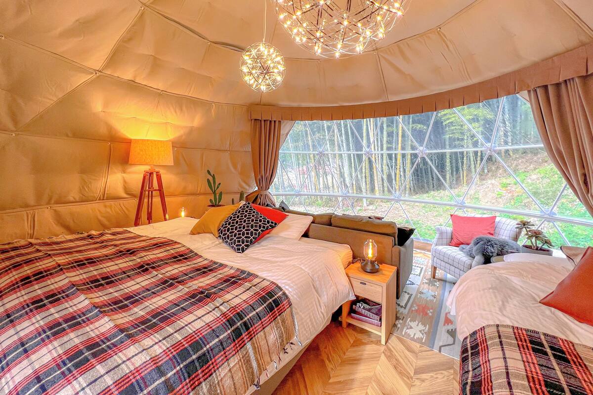 Ein gemütliches Schlafzimmer mit einem großen Bett, einem Nachttisch mit Lampe und Blick auf einen Wald durch ein großes Fenster.