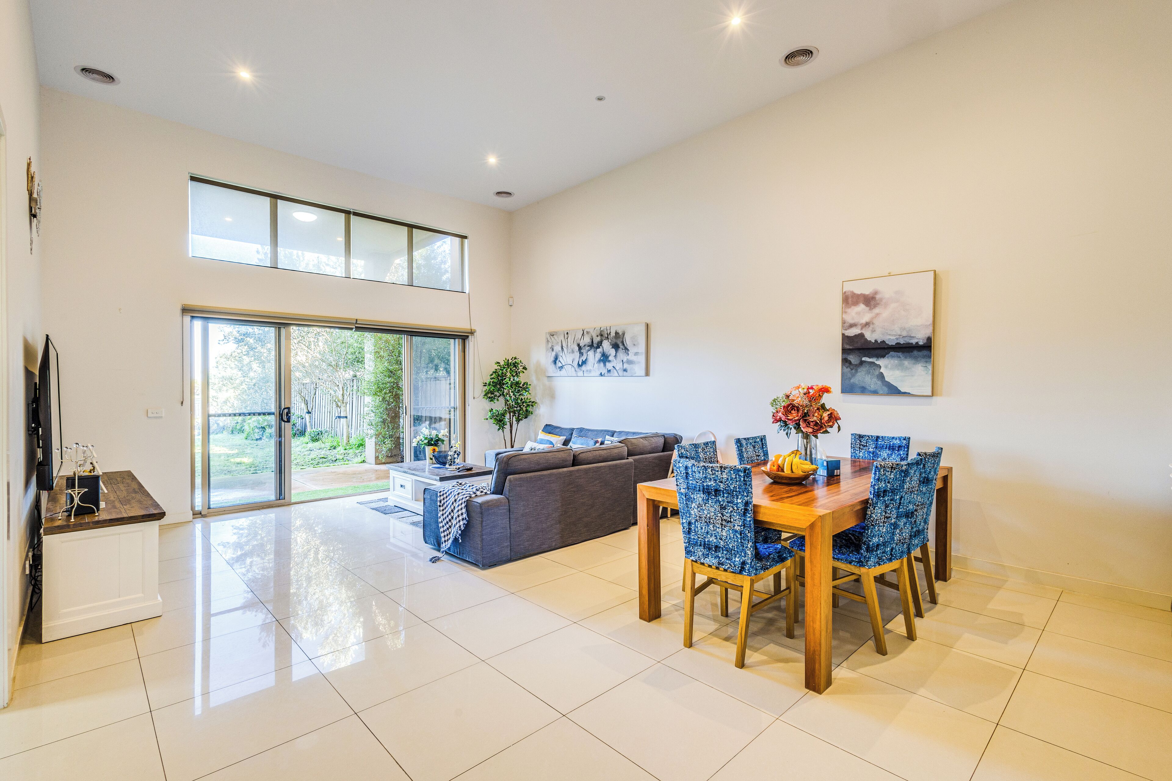 A spacious living area with a dining table, blue patterned chairs, a grey sofa, and a large window.