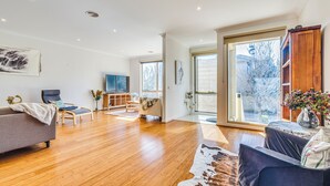 A spacious living room with hardwood flooring, a grey sofa, a blue armchair, and a large window offering a view of the outdoors. - StayAU 3BR Deluxe Family Loft Point Cook (Point Cook)