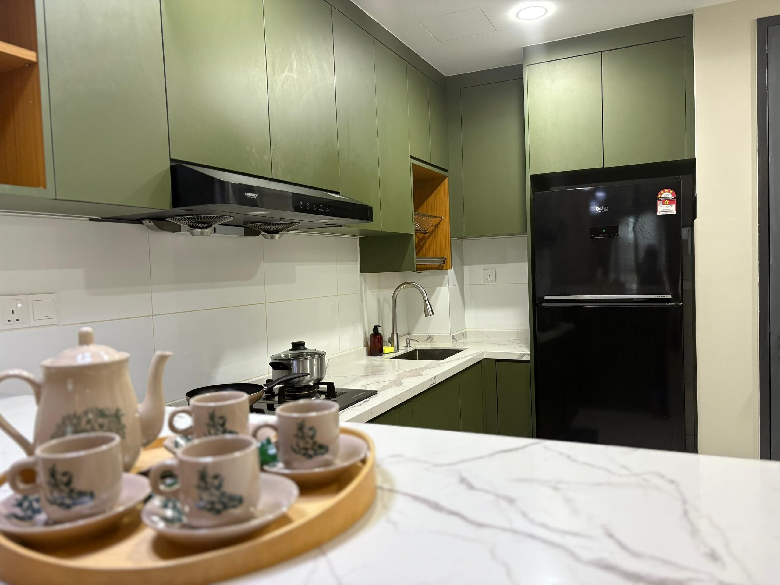 A modern kitchen with green cabinets, a black refrigerator, and a wooden tray with teapots and cups.