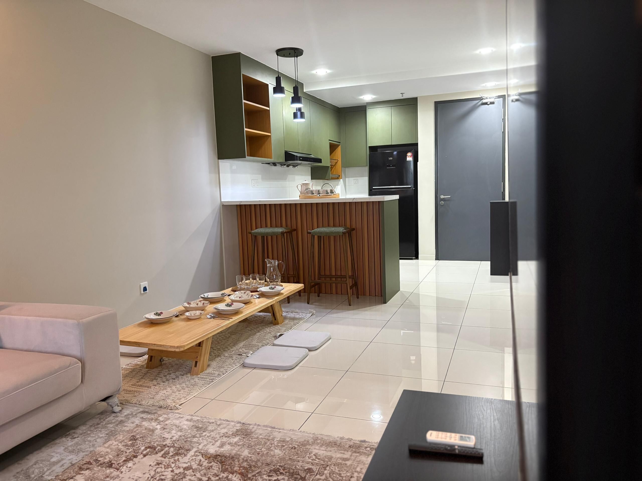 A modern living room with a sofa, a coffee table, and a dining table set for four. A kitchen with green cabinets and a black refrigerator is visible in the background.