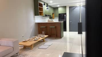 A modern living room with a sofa, a coffee table, and a dining table set for four. A kitchen with green cabinets and a black refrigerator is visible in the background.