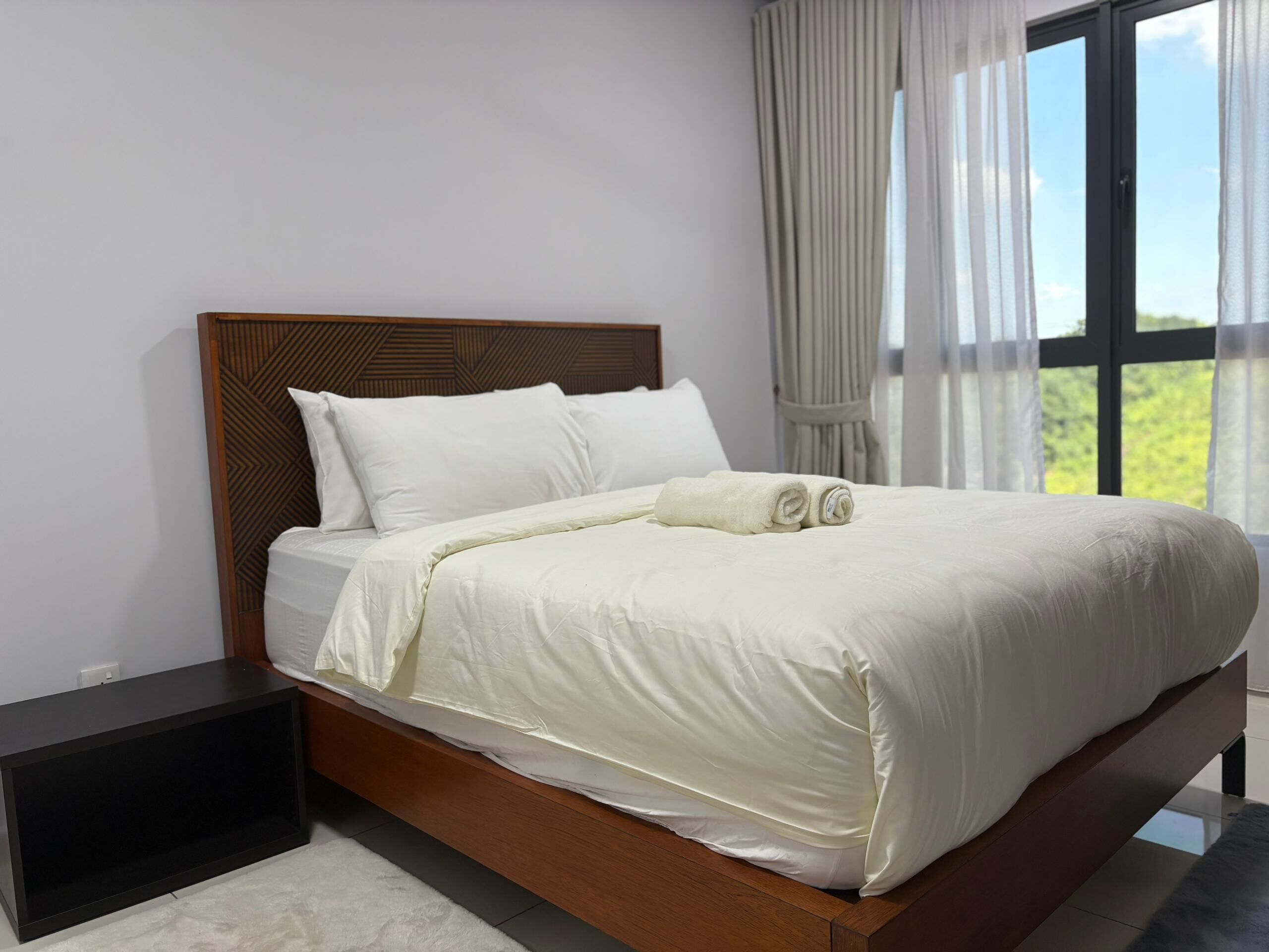 Una cama bien hecha con sábanas blancas y cabecera de madera, ubicada junto a una ventana con cortinas traslúcidas.