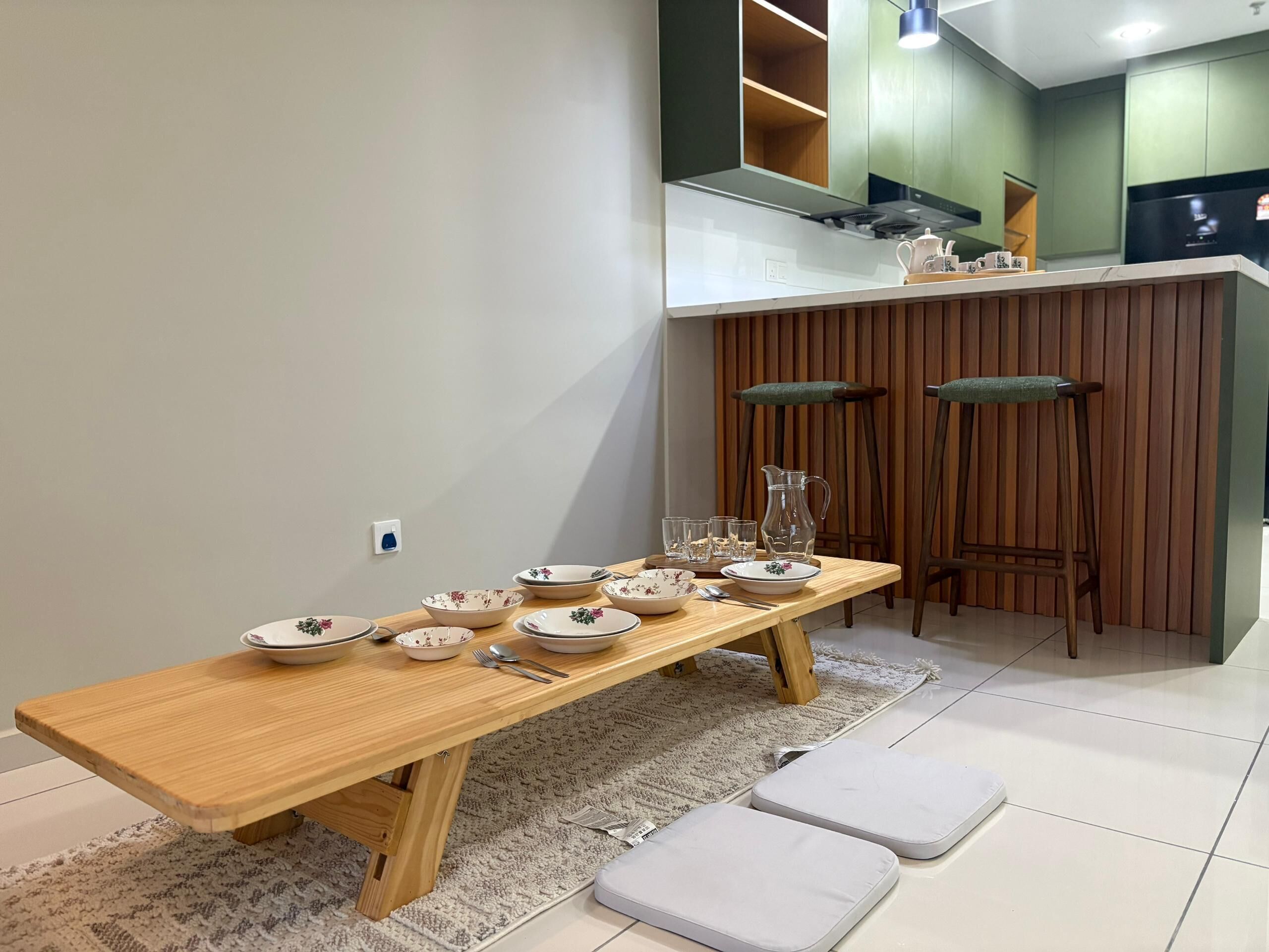Een eetgedeelte met een houten tafel gedekt voor vier personen, een keuken op de achtergrond en een zithoek met krukken.