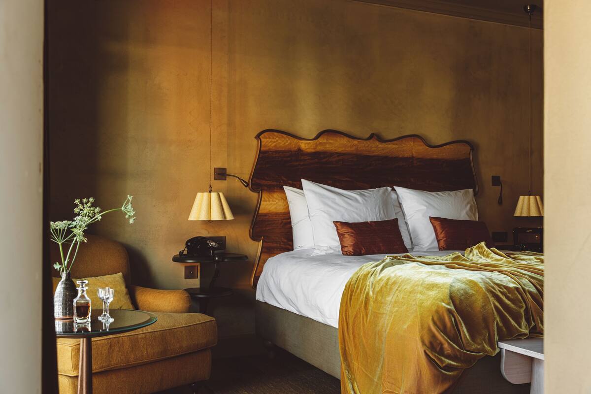 A hotel room with a large bed, a nightstand with a lamp, a small table with a vase and glasses, and a sofa.