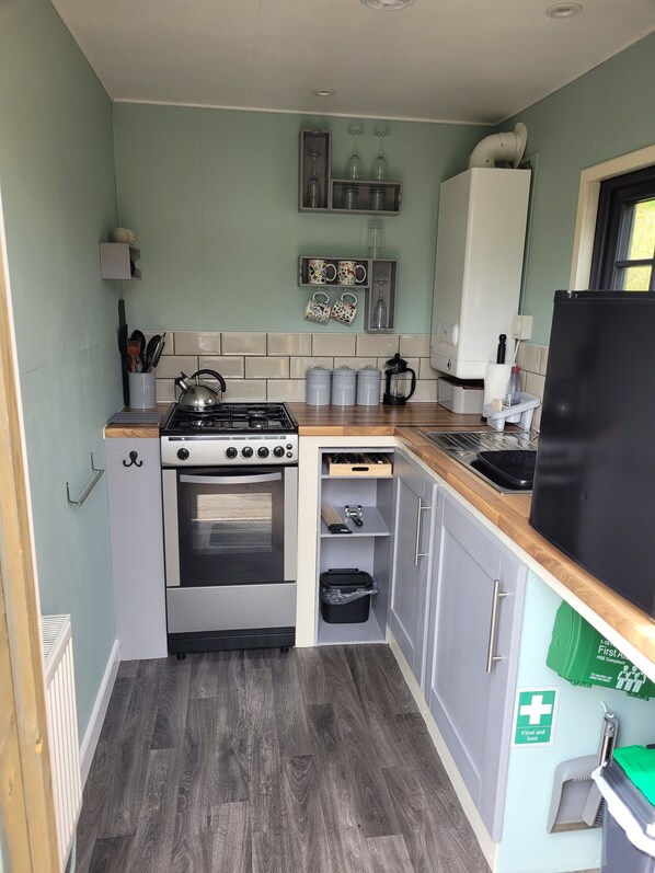 A compact kitchen with a gas stove, oven, and a built-in dishwasher. There are cabinets, a countertop, and a small window. - Go Eco Glamping (Lampeter)