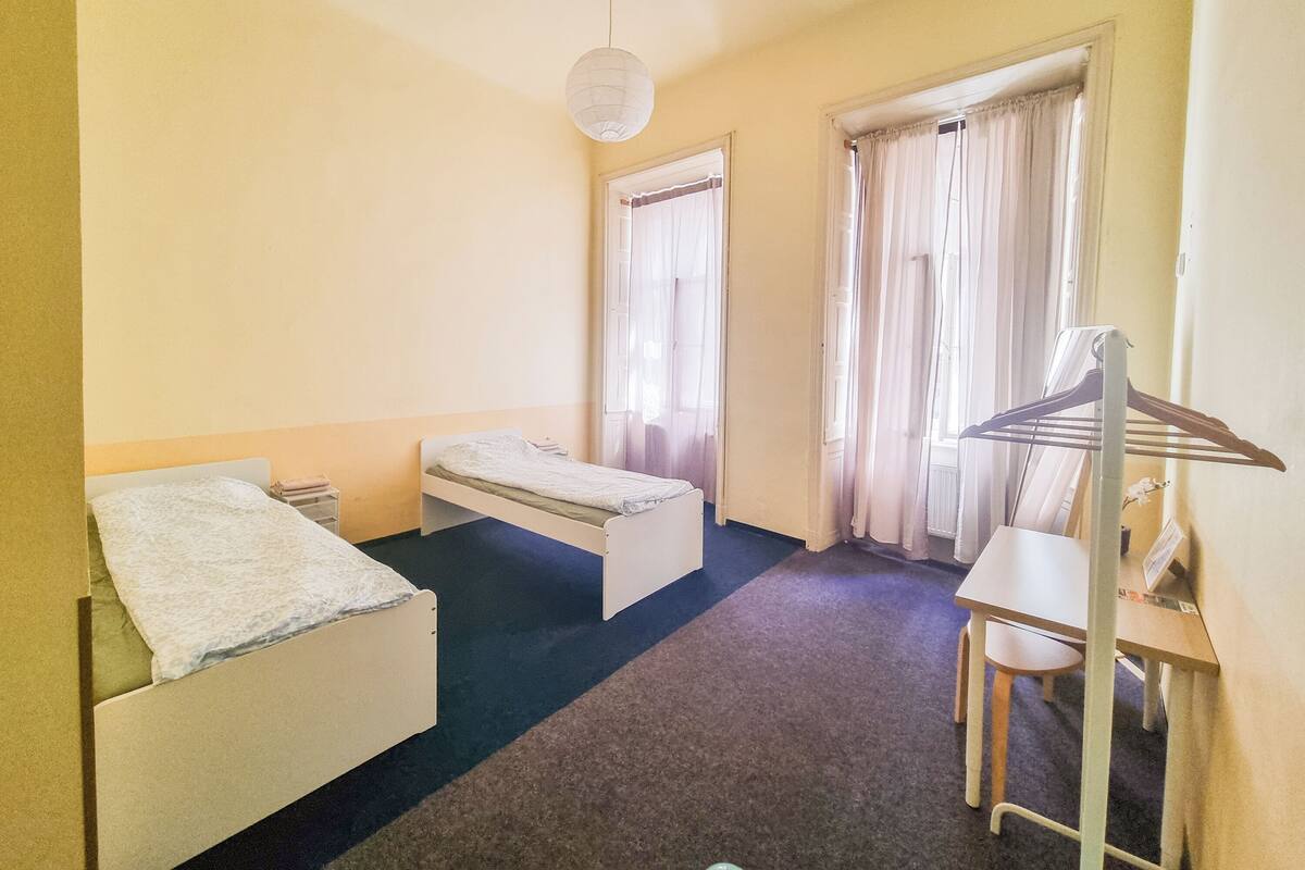 A small, simple room with two single beds, a desk, a chair, and a hanging clothes rack.