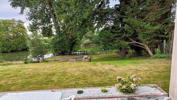 Un patio con una mesa y sillas, un arreglo floral y vistas a un área verde con árboles y una masa de agua.