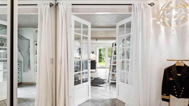 A room with a large open door leading to a balcony, a white tufted ottoman, and a black jacket on a stand. - "Mark" - 1.2km from the sea by Interhome (Græsted)